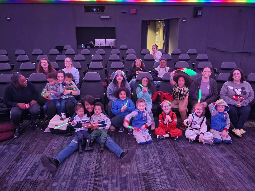 Group of adults, tweens, and kids sitting in movie theater style seats at the planetarium.